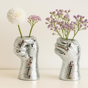 May include: Two silver mosaic fist-shaped vases, each holding flowers. One vase has a white dahlia, the other has purple baby's breath. The vases are on a white surface against a white background. The vases are decorative and unique.