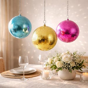Könnte beinhalten: Drei hängende Discokugeln in Blau, Gold und Pink reflektieren das Licht. Ein Esstisch ist mit Tellern, Gläsern und einem Blumengesteck gedeckt. Kerzen tragen zur festlichen Atmosphäre bei.