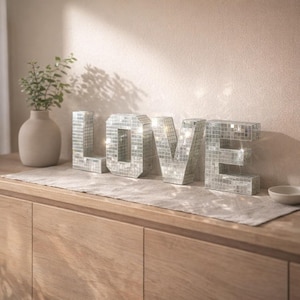 Puede incluir: Un letrero decorativo "LOVE" de mosaico plateado, compuesto por azulejos de espejo, se encuentra sobre una superficie de madera. Las letras reflejan la luz. A la izquierda hay un pequeño jarrón con vegetación. El letrero es un artículo decorativo.