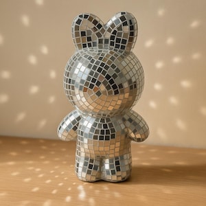 May include: A silver mosaic bunny figurine with a reflective surface. The bunny has a round body, small arms, and large ears. The figurine is on a wooden surface with light patterns on the wall.