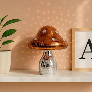 Pu&ograve; includere: Una lampada decorativa a forma di fungo con paralume a mosaico color rame e base argentata. Una pianta in vaso con foglie verdi e una stampa incorniciata con la lettera "A" sono su una mensola bianca.