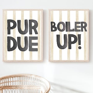 May include: Two framed prints with the words "PURDUE" and "BOILER UP!" in dark gray lettering on a cream and white striped background. The frames are light brown. A wicker chair is in the foreground.