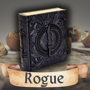 May include: A black, embossed book with a dagger design on the cover. The word "Rogue" is displayed on a banner at the bottom. The book has a decorative border and gold-colored pages. Dice are visible in the background.