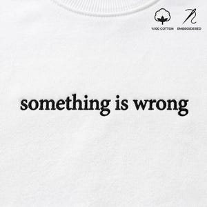 Op de afbeelding: Witte crewneck sweatshirt met de geborduurde zin "something is wrong" in zwart. De rechterbovenhoek heeft een katoenen symbool en het woord "geborduurd".