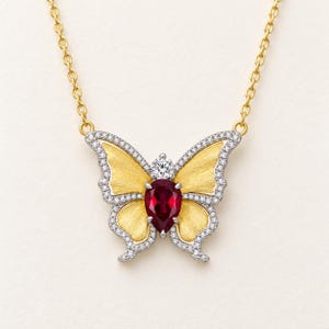 May include: A gold chain necklace featuring a butterfly pendant. The butterfly has textured yellow gold wings, a pear-shaped red gemstone in the center, a small round white gemstone above it, and a border of tiny white pavé-set stones.