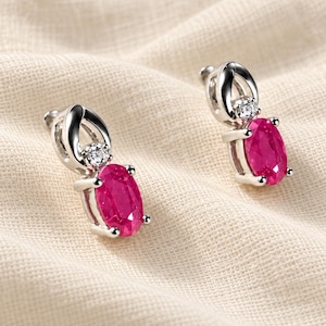 May include: A pair of silver-toned earrings featuring an oval-cut vibrant pink gemstone set in four prongs, topped with a small round clear stone held in a polished metal loop design that connects to the earring post.