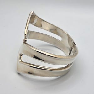 Op de afbeelding: Een zilverkleurige metalen armband met een scharnierend ontwerp. De armband heeft een dubbelbandige structuur met open ruimtes, waardoor een moderne en strakke esthetiek ontstaat. Het gepolijste oppervlak reflecteert licht en benadrukt de gladde rondingen en geometrische vorm.