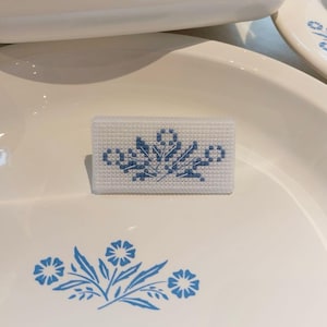 May include: A selection of white ceramic tableware featuring a blue floral pattern. The items include a rectangular casserole dish, a serving platter, and a small, cross-stitched rectangle with the same floral design.