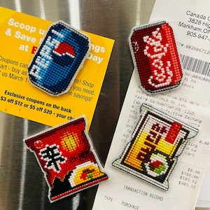 May include: Four handmade, cross-stitched magnets: a blue Pepsi can, a red Coca-Cola can, and two bags of ramen. The magnets are on a metallic surface with a yellow coupon and a receipt.