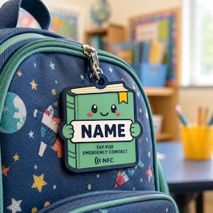 May include: A green, book-shaped silicone bag tag featuring a cartoon face, a white rectangular space labeled NAME, and the text TAP FOR EMERGENCY CONTACT with an NFC symbol, attached to a navy blue backpack with a space-themed star pattern.