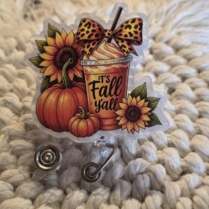 May include: An acrylic badge reel featuring a colorful illustration of two orange pumpkins, two yellow sunflowers, and a plastic cup with a straw. The cup has a leopard print bow on top and the text It's Fall y'all printed in black lettering.