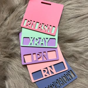 May include: A stack of colorful, rectangular badge holders in pink, mint green, lavender, peach, and blue. Each badge holder has a cutout with a profession title: RN BSN, XRAY, LPN, RN, and RESPIRATORY.