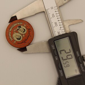 May include: An orange Sweet Georgia pomade tin with a digital caliper measuring 29.59 mm. The tin features two portraits and the text "HAIR DRESSING POMADE". The caliper is silver and black.