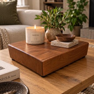 May include: A square wooden riser with a smooth finish, topped with a lit candle in a speckled beige container labeled SANTAL, a small potted green plant in a textured ceramic vase, a small wooden bowl, and two stacked square stone coasters.