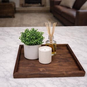 May include: A dark brown wooden tray holds a small potted plant, a white candle, and a glass bottle with reeds. The candle is white with vertical ridges. The plant has small green leaves. The tray sits on a marble surface.