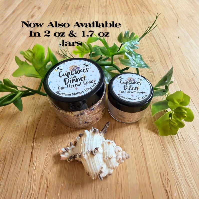 May include: Two clear plastic jars with black lids containing hermit crab food, labeled Cupcakes for Dinner, available in 2 oz and 1.7 oz sizes. A white spiral seashell sits in front of the jars, surrounded by green artificial leaves.