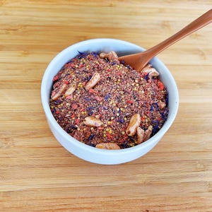 May include: A white ceramic bowl filled with a coarse, multi-colored dry spice blend containing red, yellow, and blue particles, along with several light brown, elongated seed-like pieces. A smooth, light brown wooden spoon rests inside the bowl.
