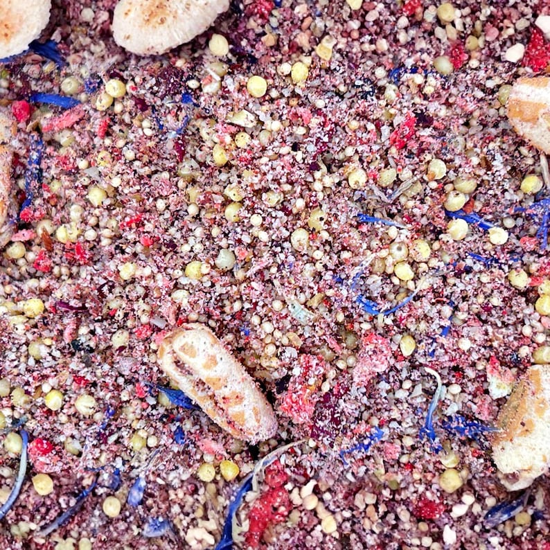 May include: A dry mixture of small, round, light yellow grains, vibrant red dried fruit pieces, and scattered bright blue flower petals. The blend includes several larger, oval-shaped, light tan textured pieces distributed throughout the surface.