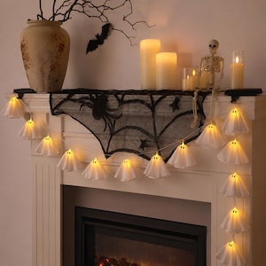 May include: A string of white ghost-shaped LED lights with black eyes, draped across a mantelpiece. The display includes a black spiderweb runner, a beige vase with bare branches, a paper bat, several pillar candles, and a small skeleton figure.