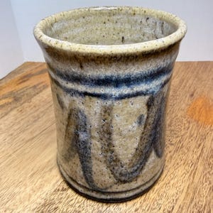 Puede incluir: Un vaso de cerámica hecho a mano con un esmalte beige y azul moteado. La taza cilíndrica presenta una banda azul oscuro y patrones ondulados verticales. El borde está ligeramente ensanchado y la pieza tiene una estética rústica y artesanal.