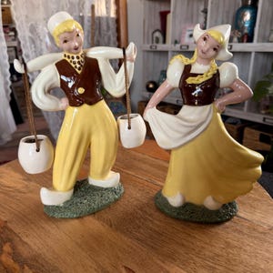 May include: Two ceramic figurines of Dutch figures. The male figure wears a white hat, shirt, brown vest, yellow pants, and wooden shoes, carrying two white pots. The female figure wears a white bonnet, white shirt, brown vest, and yellow skirt.