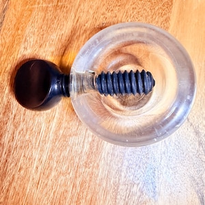 May include: A clear glass bottle stopper with a black rubber screw and a black knob. The stopper is designed to fit into a bottle opening. The stopper is on a wooden surface.