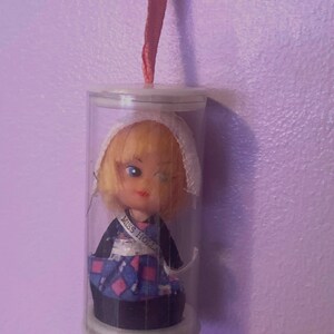 May include: A vintage doll, labeled "Miss Holland," encased in a clear plastic cylinder. The doll has blonde hair, blue eyes, and wears a traditional Dutch outfit with a blue and pink patterned skirt. A red ribbon is attached for hanging.