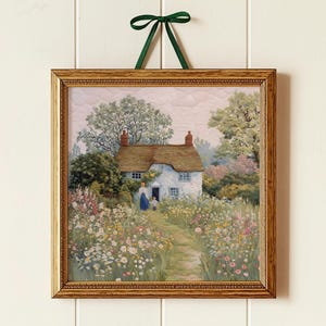 Könnte beinhalten: Ein gerahmter Druck einer Cottage-Szene mit Strohdach, umgeben von einem lebendigen Blumengarten. Der goldfarbene Rahmen wird von einem grünen Band gehalten. Das Kunstwerk zeigt einen Weg zum Cottage mit Figuren in der Ferne.