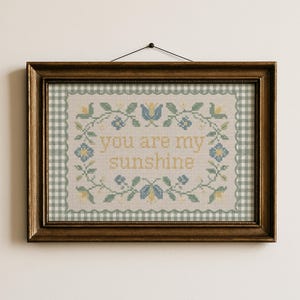 May include: A framed cross-stitch artwork with the text "you are my sunshine". The artwork features a floral border with blue and yellow accents, set against a light background. The frame is a dark brown color.