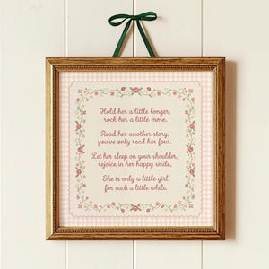 May include: A square framed wall hanging featuring a light pink gingham background and a floral border. The text reads: Hold her a little longer, rock her a little more, Read her another story, you've only read her four, Let her sleep on your shoulder, rejoice in her happy smile, She is only a little girl for such a little while. A dark green ribbon is attached to the top.
