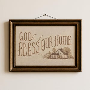 May include: A rectangular framed cross-stitch embroidery on beige fabric featuring the brown text "GOD BLESS OUR HOME" and a small house illustration. The piece is enclosed in a dark brown wooden frame with a black string for wall hanging.