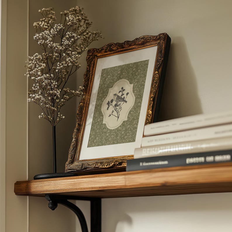 May include: A framed picture with a gold ornate frame featuring a bird and floral design. The picture is on a wooden shelf with books and a decorative plant. The frame is approximately 12 inches by 16 inches.