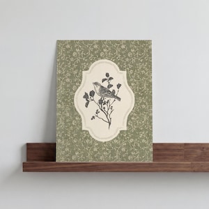 May include: A framed print featuring a detailed illustration of a bird perched on a flowering branch. The bird and branch are set against a cream-colored, decorative frame, which is set against a green floral patterned background. The print is displayed on a dark brown wooden shelf.