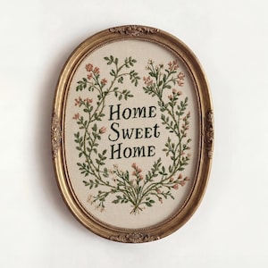 May include: An oval wall hanging featuring embroidery on beige fabric inside a decorative gold frame. The embroidery displays the text "HOME SWEET HOME" in dark green letters, surrounded by a floral wreath of green vines and small pink flowers.