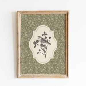 May include: Framed artwork featuring a detailed illustration of a bird perched on a flowering branch. The bird and branch are in a cream-colored, decorative shape, set against a green floral patterned background. The frame is a light brown color.