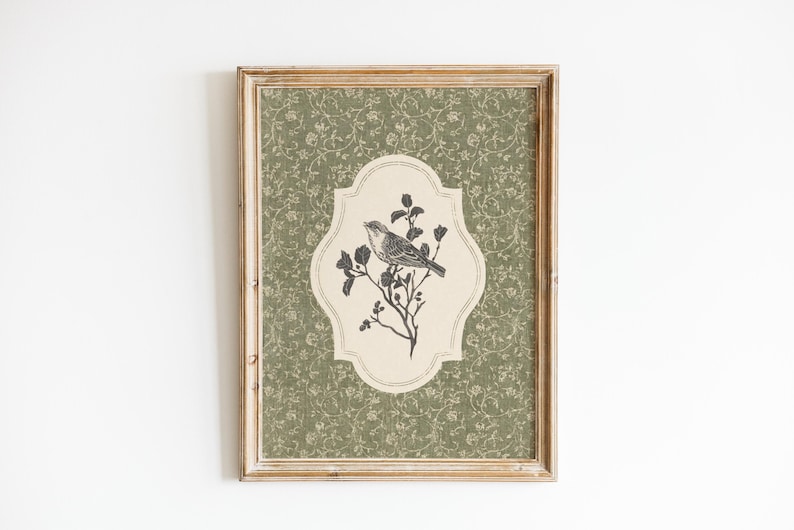 May include: Framed artwork featuring a detailed illustration of a bird perched on a flowering branch. The bird and branch are in a cream-colored, decorative shape, set against a green floral patterned background. The frame is a light brown color.