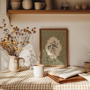 May include: A framed print featuring a bird perched on a branch, set against a green floral background. The print is displayed on a table covered with a checkered tablecloth, alongside a white mug, a book, and a vase of dried flowers.