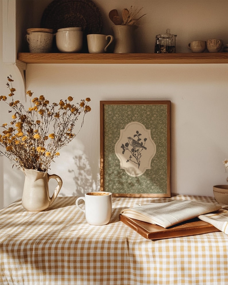 May include: A framed print featuring a bird perched on a branch, set against a green floral background. The print is displayed on a table covered with a checkered tablecloth, alongside a white mug, a book, and a vase of dried flowers.