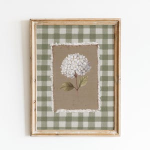 Op de afbeelding: Ingelijste bloemenkunst met een witte hortensia bloem op een bruine achtergrond. Het kunstwerk is omlijst met een verweerde gouden lijst en een groen-wit geruit patroon. Een charmant stuk voor de interieurdecoratie.