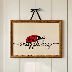 以下が含まれることがあります： 赤い水玉模様のテントウムシと筆記体で「snuggle bug」という言葉が刺繍されたアートワーク。 金色の装飾フレームに入れられ、黒いリボンから吊り下げられています。
