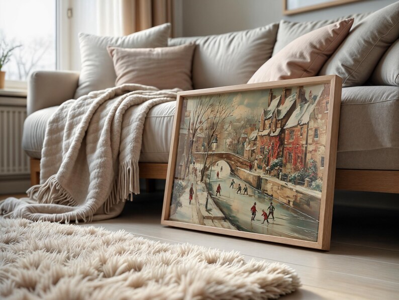 May include: A framed landscape painting of a winter scene with a bridge and figures, in a light wooden frame. The artwork is on a wooden floor. A beige rug and sofa with cushions are in the background. The painting is in a light wooden frame.