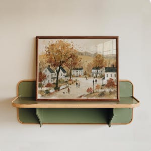 Puede incluir: Una l&aacute;mina rectangular enmarcada que muestra una escena de pueblo oto&ntilde;al estilo acuarela con casas blancas, &aacute;rboles naranjas y marrones, y figuras caminando por un sendero, expuesta sobre un estante de pared verde con bordes redondeados.