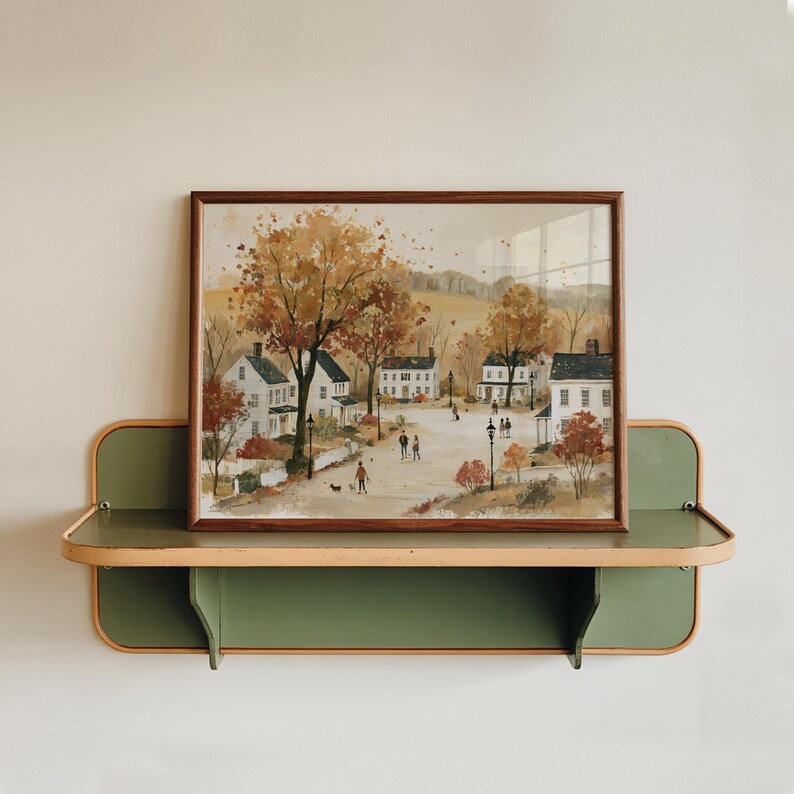 Puede incluir: Una l&aacute;mina rectangular enmarcada que muestra una escena de pueblo oto&ntilde;al estilo acuarela con casas blancas, &aacute;rboles naranjas y marrones, y figuras caminando por un sendero, expuesta sobre un estante de pared verde con bordes redondeados.