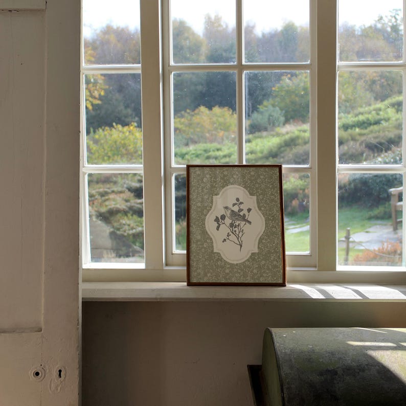 May include: A framed print featuring a bird perched on a flowering branch. The artwork is set against a light green floral patterned background, with a brown wooden frame. The print is displayed on a windowsill in front of a window.