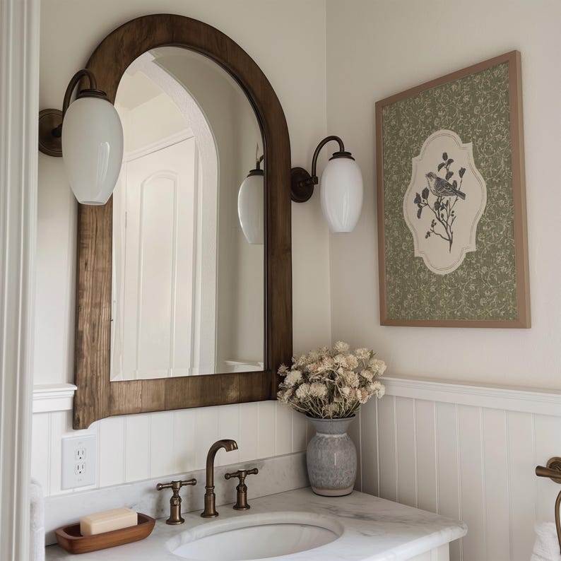 May include: A bathroom scene featuring a large, arched wooden mirror, flanked by two sconces with white globe lights. A framed artwork with a bird illustration hangs on the wall. A vase of dried flowers sits on the countertop, near the sink and faucet.