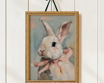 Op de afbeelding: Een ingelijst schilderij van een wit konijn met een roze strik, tegen een zachte blauwe achtergrond. Het kunstwerk zit in een gouden lijst en wordt opgehangen met een groen lint. De schilderstijl is zacht en schilderachtig.