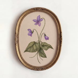 Könnte beinhalten: Ein ovaler, verzierter goldener Rahmen mit einer botanischen Illustration einer Veilchenpflanze. Das Kunstwerk zeigt drei violette Veilchenblüten mit grünen Stielen und großen, strukturierten grünen Blättern auf beigem Hintergrund.