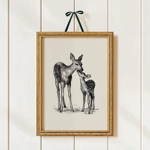 May include: A framed black and white print of two deer, one adult and one fawn, in a vintage style. The print is in a gold frame and is suspended by a green ribbon. The artwork is a nature-themed illustration.