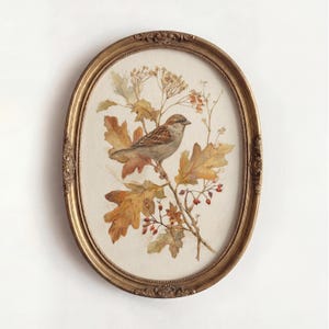 Könnte beinhalten: Ein ovaler, verzierter goldener Rahmen mit einer Aquarellillustration eines braunen Spatzen, der auf einem Ast sitzt. Der Ast ist mit goldbraunen Eichenblättern, kleinen roten Beeren und zarten Trockenblumen vor einem cremefarbenen Hintergrund verziert.