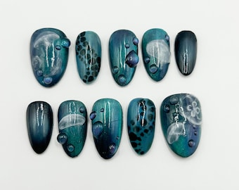 May include: A set of ten artificial nails with a teal and blue gradient design. Some nails feature a jellyfish design, while others have a bubble or spotted pattern. Each nail has a glossy finish and is adorned with small, clear, droplet-shaped embellishments.